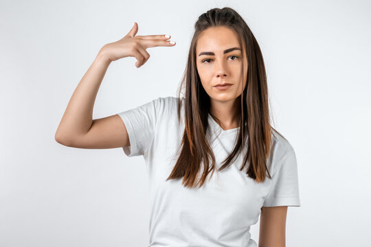 I'm fed up with this! Stylish European girl with long chestnut hair shoots in temple, tilts head, dressed in casual t shirt, demonstrates suicide gesture, isolated on white background