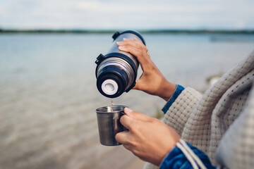 Obraz premium Tourist pours hot tea out of thermos by autumn lake. Camping, traveling. Close up of hands with cup