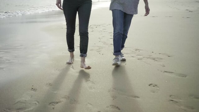 Two women walking on beach and leaving footprints in sand. Girl with barefoot and senior lady pacing along seashore and holding hands. Cropped view. Tracking shot. Leisure, and vacation concept