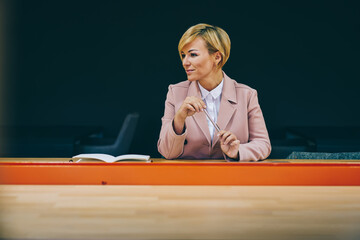Cheerful successful mature woman dressed in elegant wear smiling at looking on side.Prosperous positive female entrepreneur doing business sitting at wooden table with advertising area