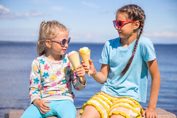 Two adorable little girls wearing sunglasses are eating ice cream on the seashore on summer vacation