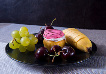 A black plate with cherries, grapes, a banana, a cupcake lies on a table in the background