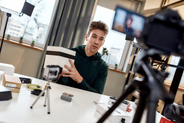 Fototapeta na wymiar Young male technology blogger recording video blog or vlog unpacking of new camera lens and other gadgets at home studio. Blogging, Work from Home concept
