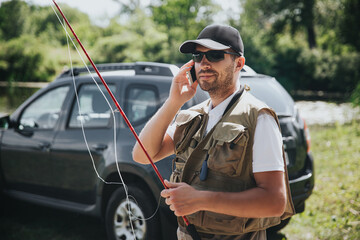 Young fisherman fishing on lake or river. Busy guy holding fishing rod and talking on smartphone. Stand alone at car during fashing activity.