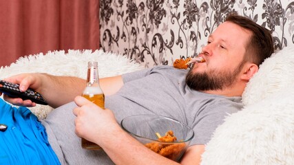 fat man drinks beer and eats unhealthy food chicken wings, bored in front of the TV outlook on the couch. The concept of malnutrition, quarantine at home, alcoholism.