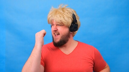 Naklejka premium freaky fat man in a wig and a pink T-shirt in his hands listens to music on headphones and dances on a blue background