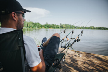 Young fisherman fishing on lake or river. Guy sitting in folding chair and looking straight forward on lake or river. Three fishing rods in front him for getting fresh fish. Water hunting.