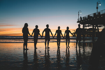 Group of young people holding each other hands standing on sea shore during sunset, male and female persons silhouettes spending time together enjoying traveling on tropical island during summer