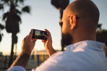 Fototapeta premium Close up rear view of young male holding in hands smartphone with modern camera for taking photos of beautiful sunset, cropped view of hipster guy shooting evening landscape on mobile telephone