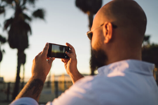 Close up rear view of young male holding in hands smartphone with modern camera for taking photos of beautiful sunset, cropped view of hipster guy shooting evening landscape on mobile telephone
