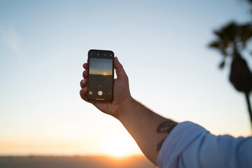 Cropped image of man's hand holding smartphone shooting video of evening sky and sundown, male using modern mobile phone camera taking photo of golden sunset  and nature during summer evening