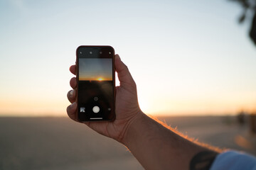 Fototapeta premium Cropped image of man's hand holding modern smartphone using application for taking evening photos of beautiful sundown, closeup view of male shooting video of sunset and skyline on telephone camera