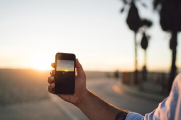 Fototapeta premium Close up view of male shooting video of sunset and skyline on smartphone camera, cropped image of man's hand holding mobile phone using app for making pictures of sundown during summer vacation