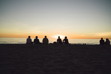 Young persons sitting on ocean beach enjoying recreation during sundown on tropical island, peoples silhouette of friends looking at beautiful nature landscape in summer evening dusk on seaside .