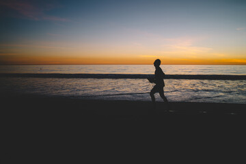 Male silhouette of jogger starting day with activity in morning dusk on ocean shoreline, man running on sandy beach in evening twilight and sunset spending summer vacation on tropical island.