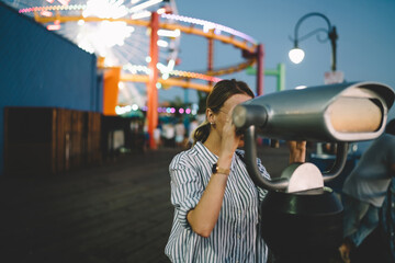 Hipster girl looking at view in coin operated optical binocular standing in amusement park shining with night lights recreating on summer vacations, female tourist resting in Santa monica pier.