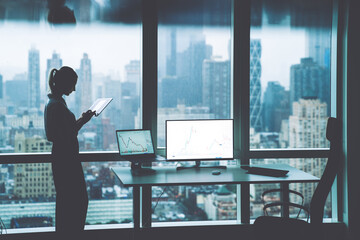 Silhouette of female person hold in hands touch pad while standing near office desktop with Mock up PC computer screen front panoramic city view in window.Trade manager workspace with financial graphs