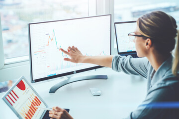 Back view of business woman sitting at desktop front PC computer with financial graphs and...