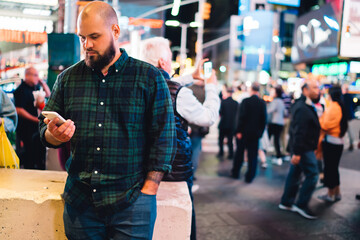 Pensive bearded man holding mobile phone reading message standing on Times square at evening,hipster guy using smartphone and 4G connection in roaming for browsing spending weekends in New York