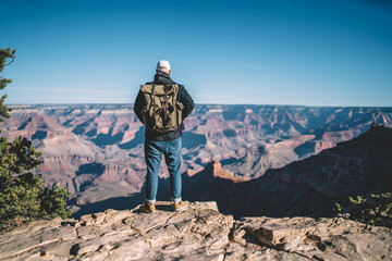 Back view of male traveler enjoying view from mountain top getting to destination of hiking tour, hipster guy with backpack standing on rocky hill looking at dry valley in Grand canyon National Park