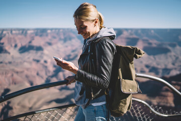 Fototapeta premium Smiling female explorer reading message on telephone satisfied with mobile connection during hiking tour,cheerful girl wanderlust using cellular chatting standing on viewpoint in Grand Canyon.