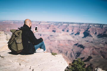 Fototapeta premium Back view of male explorer wanderlust using smartphone for shooting video for travel blog sitting on high rock in mountains during trip, millennial hipster guy tourist making selfie during hiking tour