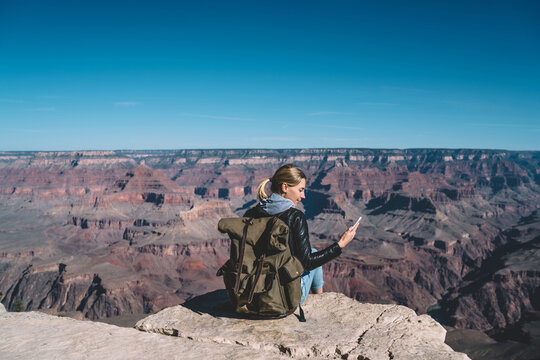 Female traveler holding smartphone checking mobile data connection sitting on mountain top in Grand Canyon National Park, hipster girl enjoying hiking with good network coverage for sending messages