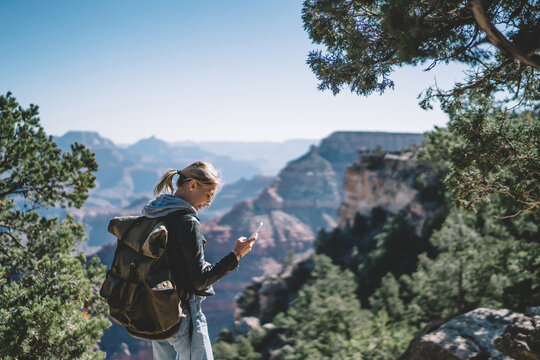 Smiling female tourist with backpack satisfied with having roaming mobile connection for communication in wild environment, hipster girl navigating during hiking tour using application on smartphone