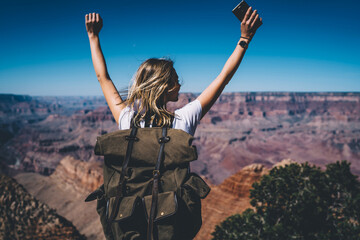 Back view of excited hipster girl standing on high mountain top feeling free raising hands up, happy female traveler with backpack celebrating achievement of reaching peak during hiking tour in USA