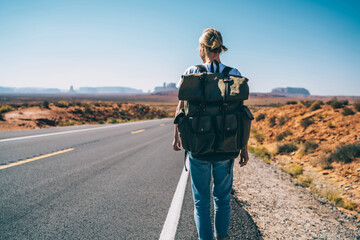 Back view of female traveler passing wild lands near asphalt road exploring southwest of USA,hipster girl enjoying active lifestyle with backpack walking on roadway having journey with hitchhiking