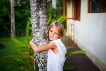 Portrait. Charming little girl standing and holding the tree, wearing white dress. Cute face. Spending time outdoor in tropical garden. Childhood concept.