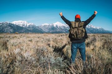 Back view of hipster guy standing with raising hands feeling excited with beautiful view of winter mountains ahead, male traveler wanderlust happy and celebrating achievement getting to destination