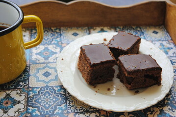 Chocolate cake slices in a white ceramic plate and cup of Turkish coffee in a yellow enamel mug served on the wooden tray with azulejos motif. Dessert time.  