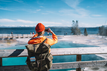 Back view of female tourist with backpack using smartphone for taking picture of hot springs,...