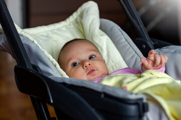 Photo of cute newborn baby sitting in stroller at home.Happy smiling baby girl in a stroller indoors.Side view image of baby girl in pink bodysuit sitting in modern stroller.Little kid in a pushchair.
