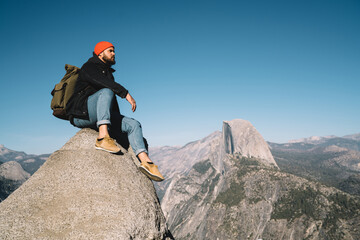 Serious male traveler in red hat sitting on high top of rock looking at valley scenery exploring wanderlust getaway, hipster guy tourist enjoying nature and extreme climbing in mountain landscape
