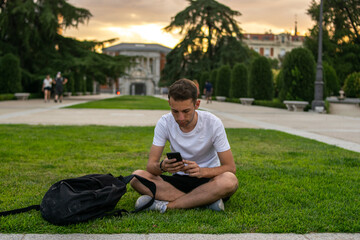 Fototapeta premium Photo of a young and attractive man with a white t-shirt sitting on the grass checking his phone