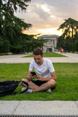 Fototapeta premium Photo of a young and attractive man with a white t-shirt sitting on the grass checking his phone