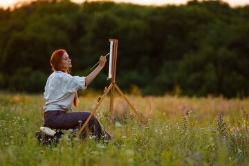  a girl artist with long red hair draws on an easel with a brush against the background of the sunset