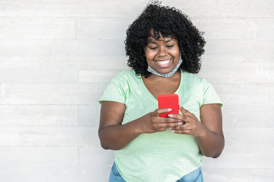 Happy black girl using smartphone outdoor - Young curvy african female having fun watching videos on mobile phone while wearing face protective mask - Coronavirus lifestyle concept - Focus on face