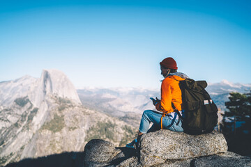 Obraz premium Female tourist with backpack sitting on mountain peak sending text messages on smartphone via good data connection,hipster girl enjoying beautiful landscape of high rock cliffs reading mail on mobile