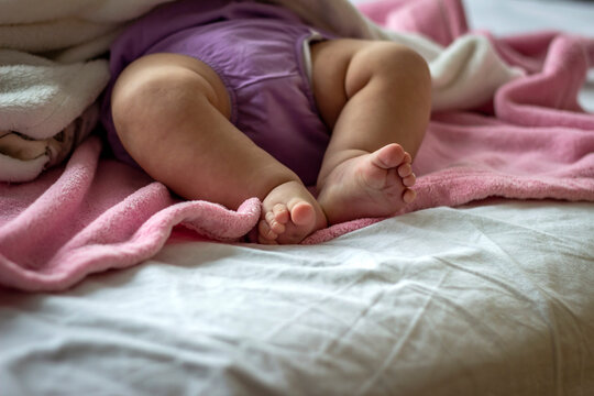 Close-up of unrecognizable cute baby girl feet lying in bed, innocence concept.Side view photo of a baby legs and bottom in diaper pants wrapped in soft pink blanket on white bed background,copy space
