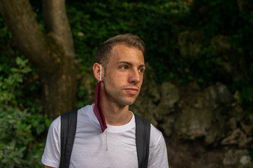 Photo of a young and attractive man with a backpack wearing a reusable face mask hanging in his ear. 