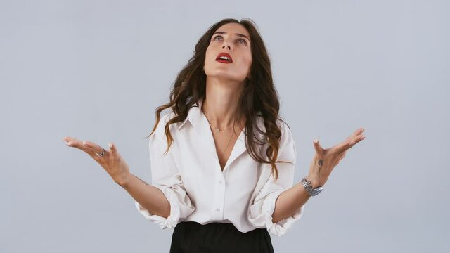 Model in white shirt is raising hands up like asking why, grabbing her head, looking disappointed and upset. Posing on gray background. Close up