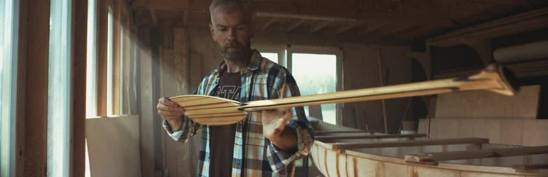 Mid 50s Caucasian male examining a hand made canoe paddle in his workshop. Boat making hobby, small business owner. Shot on RED Dragon