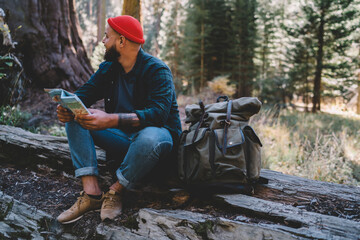 Male traveler enjoying recreating time holding map for search destination during hiking tour to discover wild nature, guy wanderlust looking around for breathtaking in spring forests sitting on log
