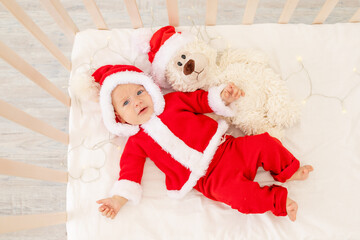 Christmas photo of a baby in a Santa costume lying in a crib at home with a toy in a Santa Claus hat, top view, happy new year