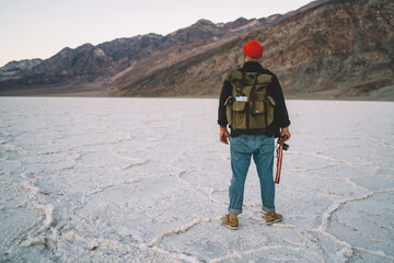 Back view of male traveler having adventure tracking touristic route in Death Valley with arid surface,photographer with backpack exploring landscape of Badwater basin with lowlands and rocks.