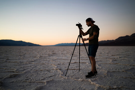 Young male with camera shooting video of wild nature of death valley working at evening dusk with modern equipment, professional photographer making photos of sunset during trip in Badwater basin