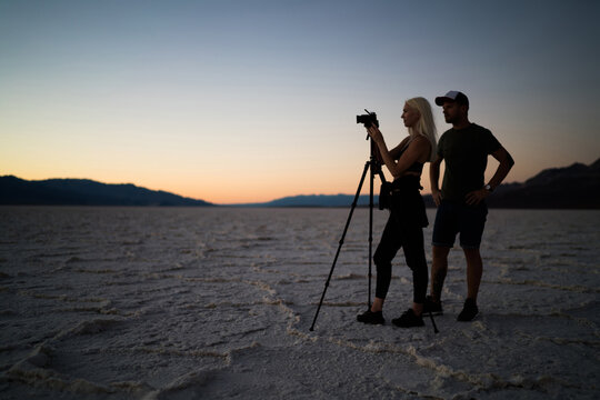 Young male and female photographers shooting video of beautiful landscape scenery, skilled caucasian woman and man taking photos of Badwater basin using modern camera and tripod for evening pictures
